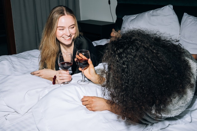 Two girls clinking red wine glasses on a bed